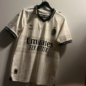 AC Milan vit bortatröja Puma - AC Milan bortatröja i vit med svarta detaljer och subtilt mönster. Puma-logga på bröstet, klubbmärke och sponsortryck. Svart mönstrad krage och ärmslut. Tillverkad i lätt och ventilerande material, perfekt för match eller träning.