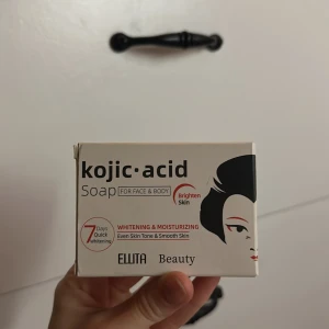 Kojic Acid Soap från Ellita Beauty - Kojic Acid Soap för ansikte och kropp från Ellita Beauty. Tvålen är orange och kommer i en vit förpackning med svart och röd design. Den utlovar whitening och återfuktning, jämn hudton och mjuk hud. Innehåller kojicsyra och levereras med ett nät för skum.