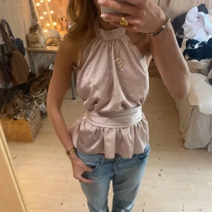 Champagnefärgad omlottopp i satin - Säljer en champagnefärgad halterneck topp i satin med hög hals och bred knytning i midjan. Toppen är ärmlös och har en snygg peplum-effekt nedtill. Lappen är bortklippt men är från french connection i storlek M❣️säljer då den var för stor på mig i S