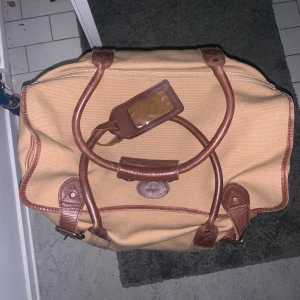 Beige resväska från Melvill & Moon - Snygg och rymlig resväska från Melvill & Moon i beige canvas med bruna skinnhandtag och detaljer. Väskan har ett randigt innerfoder i beige, grönt och rött, samt en praktisk dragkedja. Perfekt för dig som vill resa med stil och ha plats för allt du behöver.