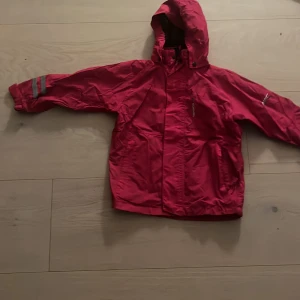 Röd regnjacka från Tenson med huva - Röd regnjacka från Tenson med huva och reflexdetaljer på ärmarna. Jackan har dragkedja framtill, två stora fickor och meshfoder på insidan. Perfekt för blöta dagar och enkel att slänga på sig när det regnar. Storlek 120