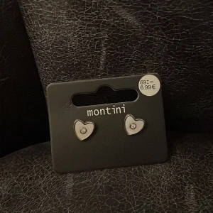 Hjärtformade örhängen från Montini - Säljer ett par söta örhängen från ur och Penn i form av hjärtan. De är silverfärgade med en liten glittrande sten i mitten som ger extra bling. Perfekta för dig som gillar stilrena och romantiska accessoarer.
