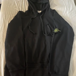 Svart hoodie från Stone Island - Svart hoodie från Stone Island med klassisk logotyp-patch på vänster ärm. Tröjan har huva med snörning och en stor magficka. Materialet är mjuk bomull och passformen är normal. Perfekt för dig som gillar streetwear och vill ha något stilrent och bekvämt.