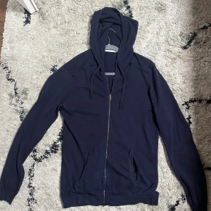 Mörklblå kashmir/ull zip-hoodie - Mörkblå hoodie med dragkedja. Tröjan har huva med snörning, långa ärmar och två fickor framtill. Materialet är kashmir och ull blandning. Enkel och stilren design som funkar till det mesta.