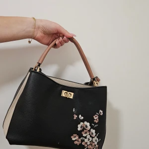 Svart handväska med broderade blommor - Snygg svart handväska i skinnimitation med beige sidor och brunt handtag. Väskan har guldfärgade metalldetaljer och ett vackert broderat blommönster i vitt och rosa på framsidan. Perfekt storlek för att få plats med allt du behöver.