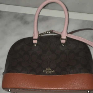 Coach handväska med rosa handtag - Snygg handväska från Coach med klassiskt logomönster i mörkbrunt och svart. Väskan har ljusrosa handtag och axelrem samt en brun botten i skinn. Perfekt storlek för det viktigaste. Köpte för 3200 kr. Säljer för 2800. Köptes för ett år sedan men aldrig använd och i mycket bra skick 💖