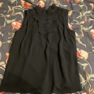 Svart blus med spetsdetaljer - Säljer en svart blus med hög hals och snygga spetsdetaljer framtill. Blusen är ärmlös och har en lätt, flowy passform. Perfekt för dig som gillar stilrena plagg med en twist. Stängs med knapp i nacken och har en diskret volangkant vid halsen.