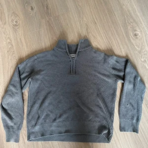 Grå stickad half zip tröja - Snygg grå stickad tröja med ribbad struktur och half zip-dragkedja i halsen. Perfekt för lager-på-lager och chill stil. Långärmad och med hög krage som ger extra edge till outfiten.