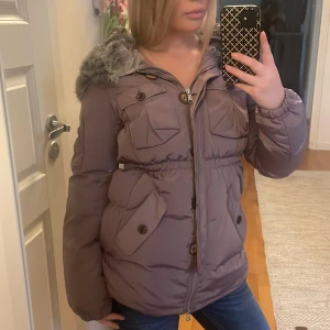 pufferjacka Odd Molly med päls - Säljer en lila pufferjacka från Odd Molly med stor huva och grå fuskpäls. Jackan har flera fickor med knappar, dragkedja och markerad midja. Den säljs inte längre. 