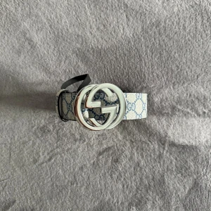 Gucci bälte  - Säljer ett Gucci GG Supreme bälte med klassiskt GG-mönster i blått på beige bakgrund. Bältet har en stor rund silverfärgad spänne med Gucci-logga och är tillverkat i skinn med canvas-ytan. Perfekt för att lyfta din outfit med en lyxig touch.