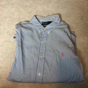 Randig blå skjorta från Polo Ralph Lauren - Slim fit skjorta från Polo Ralph Lauren i storlek L. Skjortan har blå och vita vertikala ränder, klassisk krage med knappar och den ikoniska rosa broderade loggan på bröstet. Tillverkad i mjuk bomull och har långa ärmar med knappslå.