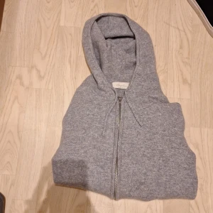 Grå kashmir hoodie från Sayless - Grå hoodie från Sayless i mjuk kashmir. Tröjan har huva, snörning och tvåvägsdragkedja framtill. Perfekt för dig som gillar chill och stilren look. Ribbstickade muddar och klassisk passform. Designad i Sverige.