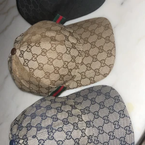 Gucci keps med GG-mönster och ränder - Snygg Gucci keps med klassiskt GG-mönster över hela kepsen och ikoniska gröna och röda ränder på sidan. Finns i beige med brunt mönster och i beige med marinblått mönster samt helsvart med svarta GG-loggor. Justerbar baktill och tillverkad i slitstark bomull.