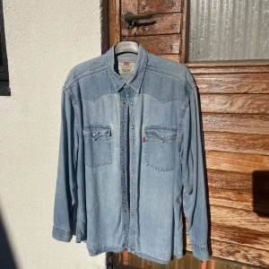 Levi's ljusblå jeansskjorta - Ljusblå jeansskjorta från Levi's med klassisk westernstil, tryckknappar och två bröstfickor med lock. Skjortan har lång ärm och är tillverkad i bomull. Perfekt för en avslappnad och cool look.