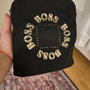 Svart axelväska från Hugo Boss - Svart axelväska från Hugo Boss med coolt cirkeltryck i guld på framsidan. Väskan har en bred, vit och grå axelrem som ger en sportig vibe. Perfekt storlek för mobilen och plånboken. Materialet är slitstark polyester och väskan stängs med dragkedja.