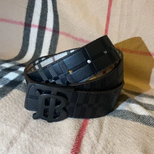 Svart rutigt Burberry bälte i skinn - Snyggt svart bälte från Burberry med klassiskt rutigt mönster och en stilren matt svart spänne med Burberrys ikoniska 'B'-logga. Bältet är tillverkat i skinn och har en modern, elegant look som passar till jeans eller kostymbyxor. Tvåsidigt storlek 105