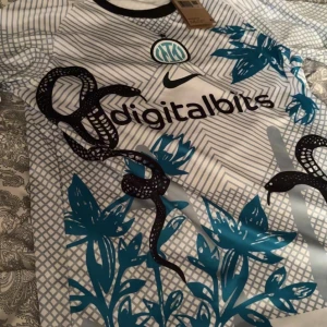 Inter Milan fotbollströja Nike blå/vit - Inter Milan fotbollströja med digitalbits-tryck, Nike-logga och klubbmärke. Tröjan är vit med tunna grå ränder, blå blommor och svarta ormmotiv. Rund halsringning med svart kant. Tillverkad i lätt och ventilerande material.