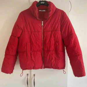Röd pufferjacka med dragkedja - Snygg röd pufferjacka med hög krage och silverfärgad dragkedja framtill. Jackan har två fickor på sidorna och justerbara snören i nederkant. Små fläckar vid kragen, knappt synliga. Se bild.
