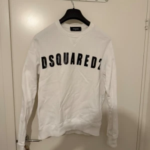 Vit sweatshirt från Dsquared2 - Säljer en vit sweatshirt från Dsquared2 med svart tryck över bröstet. Tröjan har rund hals, långa ärmar och är tillverkad i mjuk bomull. Perfekt för dig som gillar stilrena och trendiga märkesplagg. Äkta såklart 