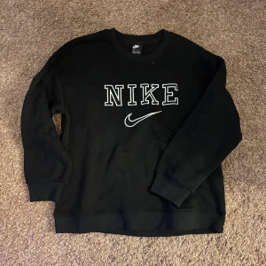 Svart Nike sweatshirt med logga - Svart sweatshirt från Nike med stor vit broderad logga och swoosh på bröstet. Klassisk rund halsringning och ribbade muddar vid ärmslut och nederkant. Perfekt för en sportig och avslappnad stil.