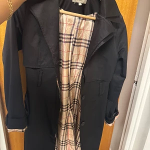 Svart trenchcoat med rutigt foder - Stilren svart trenchcoat med klassiskt rutigt foder i beige, rött och svart. 