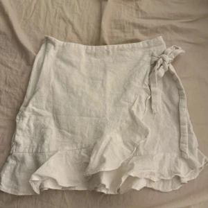 Vit omlottkjol med volang från SHEIN - Supersöt vit omlottkjol från SHEIN i storlek XS. Kjolen har volangkant nertill och knyts i sidan för en extra snygg detalj. Perfekt för en fräsch och somrig look. Materialet känns lätt och luftigt.