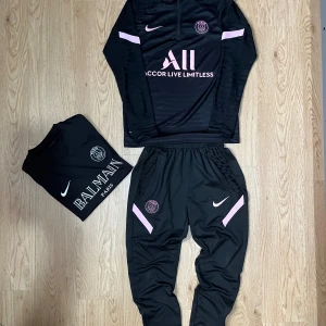 PSG x Nike tracksuit + Balmain t-shirt – storlek S/M - Snygg PSG x Nike tracksuit i svart med rosa detaljer, tillsammans med en svart Balmain Paris t-shirt. Båda i storlek S/M och i mycket bra skick – nästan som nya! Perfekt outfit för dig som gillar stilren sportlook med exklusiv touch.  ✅ Storlek: S/M ✅ Färg: Svart med rosa detaljer ✅ Märken: Nike, PSG, Balmain ✅ Skick: Mycket bra 📦 Snabb och noggrann leverans  Tracksuiten består av långärmad topp med dragkedja och matchande byxor. Balmain-t-shirten är bekväm och sitter snyggt – perfekt till både 