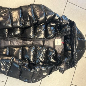 Svart glansig dunjacka från Moncler - Säljer en svart, glansig dunjacka från Moncler med klassisk puffer-design. Jackan har huva, dragkedja framtill och en ficka med Moncler-logga på ärmen. Perfekt för kalla dagar och riktigt snygg streetstil. Nfc funkar!