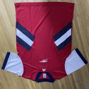 Arsenal retro fotbollströja röd/blå - Säljer en klassisk Arsenal retro fotbollströja från Score Draw i rött med vita korta ärmar och mörkblå detaljer. Tröjan har det ikoniska kanon-emblemet på bröstet och snygga blåvita ränder på axlarna. Tillverkad i polyester för skön känsla på planen.