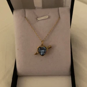 Halsband med blå hjärtberlock och pil - Gulligt halsband med tunn guldfärgad kedja och ett hänge i form av ett blått hjärta med en pil genom. Hjärtat är facetterat och gnistrar snyggt, inramat i guldton. Perfekt accessoar för dig som gillar romantiska detaljer. Rostfri