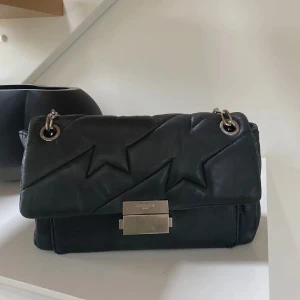 Svart  handväska från  Zadig & Voltaire - Snygg svart handväska från Zadig & Voltaire i mjukt skinn med quiltad design och coola blixtdetaljer. Väskan har silverfärgade metalldetaljer, kedjeaxelrem och ett emblem med vingar på baksidan. Perfekt storlek för det viktigaste och med medföljande dustbag.