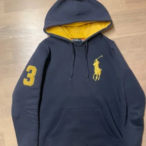 Svart gul hoodie Polo Ralph Lauren - Svart hoodie från Polo Ralph Lauren med stor gul broderad logga på bröstet och siffran 3 på ärmen. Insidan av huvan är gul, och tröjan har känguruficka och snörning i halsen. Inga skavanker eller andra slitningar. Modellen är 183cm och 69kg)
