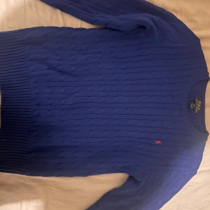 Blå kabelstickad tröja Polo Ralph Lauren - Tröjan passar bra om du är storlek S eftersom den är på barn storlek. Kabelstickad blå tröja från Polo Ralph Lauren med rund halsringning och klassisk röd broderad logga på bröstet. Tröjan har långa ärmar och ribbade muddar vid ärmslut och nederkant. Perfekt för lager-på-lager under höst och vinter💕.