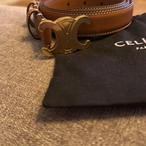 Brunt skärp med guldspänne från Celine - Säljer ett snyggt brunt bälte från Celine Paris i skinn med dekorativa vita sömmar längs kanterna. Bältet har ett ikoniskt guldspänne med Celine-logga och stilren design. Perfekt accessoar för att lyfta din outfit med exklusiv känsla.