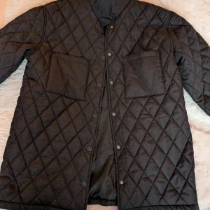 Svart quiltad jacka från KappAhl - Säljer en svart quiltad jacka från KappAhl i storlek 170. Jackan har två stora bröstfickor, tryckknappar framtill och ett stilrent rutmönster. Perfekt för dig som gillar en clean och modern look.