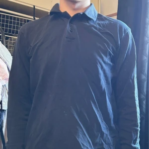 Mörkblå långärmad pikétröja - Säljer en stilren mörkblå pikétröja med lång ärm och klassisk krage. Tröjan har knäppning framtill och är gjord i mjuk bomull, perfekt för en clean och enkel look. Passar dig som gillar basic men ändå vill ha något snyggt.