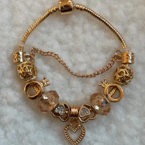 Guldigt armband med hjärtberlocker - Snyggt guldigt armband med flera berlocker, bland annat hjärtan med glittriga stenar och kronor. Armbandet har detaljer i beige och transparent samt en kedja för extra stil. Perfekt för dig som gillar unika smycken med charmiga detaljer.