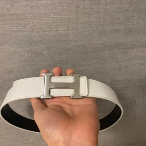 Vit herms bälte - Säljer ett stilrent vitt bälte i skinn med ikoniskt H-spänne i silverfärgad metall. Bältet har en slät och bred design med snygga sömmar och svart insida. Perfekt för dig som vill ha en lyxig accessoar med statement.