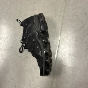 Nike Air Vapormax Plus Svarta 42 - Nike Air Vapormax Plus i helsvart färg med chunky, genomskinlig sula och snygga vågiga detaljer på sidan. Skorna har snörning och en modern, sportig look som sticker ut. Perfekt för dig som gillar streetwear och vill ha något som syns.