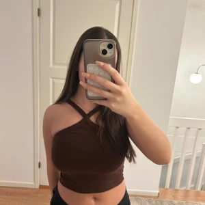 Brun ribbad croppad topp Gina Tricot - Säljer en brun croppad topp från Gina Tricot i ribbat material. Toppen har en snygg omlott-detalj med korsade axelband och är ärmlös. Perfekt för dig som gillar trendiga och stilrena plagg med en twist.