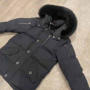 Mörkblå pufferjacka från Moose Knuckles - Säljer en mörkblå pufferjacka från Moose Knuckles med fet päls på huvan, ikonisk logga på ärmen och coola detaljer som dragkedjor och stora knappar. Jackan har flera fickor och en schysst passform som funkar perfekt när det är kallt ute.