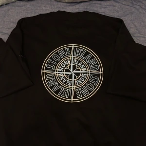 Svart Stone Island sweatshirt XL - Svart sweatshirt från Stone Island i storlek XL med stort grafiskt tryck på ryggen och klassisk logotyp på bröstet. Tröjan har ribbade muddar och den ikoniska patchen på ärmen. Perfekt för dig som gillar streetwear och vill sticka ut.
