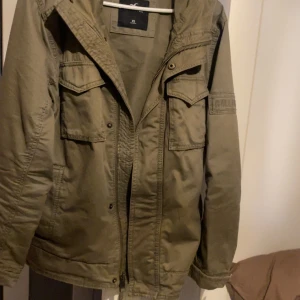 Olivgrön parkajacka Hollister XS - Snygg olivgrön parkajacka från Hollister med huva och flera praktiska fickor framtill. Jackan har dragkedja och tryckknappar, samt en patch med Hollister-logga på ärmen. Perfekt för dig som gillar en avslappnad och cool stil.