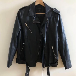 Svart skinnjacka med dragkedjor - Snygg svart skinnjacka från zara med klassisk bikerstil. Jackan har flera silvriga dragkedjor, axelklaffar och bälte i midjan. Perfekt för dig som vill ha en edgy look med stilrena detaljer och cool passform.