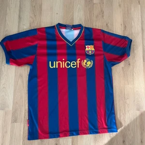 Barcelona Messi 10 fotbollströja - Säljer en klassisk Barcelona fotbollströja med Messi och nummer 10 på ryggen. Tröjan har blå och röda vertikala ränder, korta ärmar och är tillverkad i ett lätt, syntetiskt material som andas. Perfekt för dig som älskar fotboll och Messi.