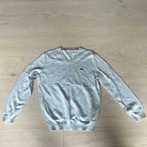  Grå v-ringad tröja från Lacoste, barn  - Snygg ljusgrå v-ringad tröja från Lacoste med klassisk krokodil-logga på bröstet. Tröjan är i mjuk bomull, har ribbade muddar och lång ärm. Perfekt för lager-på-lager och enkel att matcha med jeans eller kjol. Aldrig använd. 