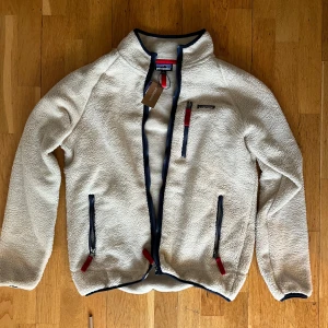 Patagonia Fleece strl L - Säljer en beige fleece från Patagonia i storlek L. Jackan har mjukt teddyfoder, tre fickor med dragkedja och snygga detaljer i marinblått och rött. Perfekt för kyliga dagar och har en chill vibe med hög krage och klassisk Patagonia-logga på bröstet. Storlek L.