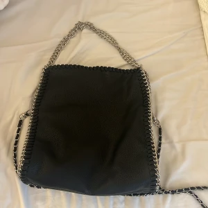 Svart axelväska med kedjedetaljer - Säljer en svart axelväska i skinnimitation med grova silverfärgade kedjor som axelrem och längs kanterna. Väskan har en rymlig insida med innerfack och dragkedja. Perfekt för dig som gillar en edgy och stilren look.