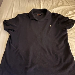 Svart piké med broderad detalj - Svart pikétröja från morris med klassisk krage och korta ärmar. På bröstet finns en liten broderad logga i vit . Materialet är mjukt och känns som bomull, perfekt för en clean och enkel look.