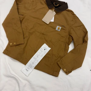 Brun Carhartt workwear jacka S - Snygg brun workwear jacka från Carhartt med klassisk krage i mörkare brun manchester. Jackan har dragkedja, bröstficka med logga och guldiga knappar vid ärmslut och midja. Tillverkad i slitstark bomull, perfekt för dig som gillar streetwear med utility-vibe. Kvitto finns. Modellen är 179 cm 65kg. Storleken är S. Möts i Sthlm eller skickar samma dag/dagen efter